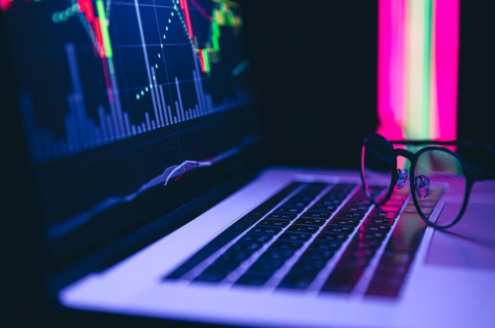 A laptop displaying colorful stock market graphs with glasses in the foreground